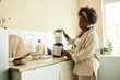 © Seventyfour - Side view of curly Black woman using food blender mixing ingredients for milk smoothie while cooking healthy breakfast at beige sunlit kitchen, copy space
