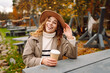 © maxbelchenko - A young woman in a stylish hat looks joyful while holding a warm beverage at an outdoor cafe. Natural outdoor scenery at the park.