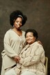 © Seventyfour - Vertical portrait of smiling African American senior woman sitting in chair hugging young daughter in beige clothes, while looking at camera on brown background in studio