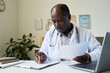 © pressmaster - Senior African American doctor reading patient records in modern, well-lit medical office focusing on healthcare management and accuracy enhancing patient care