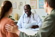 © pressmaster - Smiling doctor holding tablet while interacting with patients in consultation room. Relaxed atmosphere with plants and medical equipment visible in the background