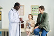 © pressmaster - Child sitting on exam table holding teddy bear while doctor talks to parent