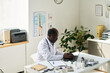 © pressmaster - African American doctor sitting at desk in bright medical office, using computer and writing, surrounded by plant and office equipment