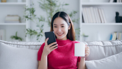 Attractive smiling young Asian woman using mobile phone while sitting on the sofa at home