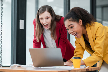 Wall Mural - Two enthusiastic businesswomen in professional attire working together on a laptop in a contemporary office setting.