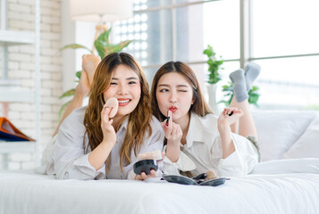 Two happy beautiful Asian female smile lying relaxed on a white bedroom, putting makeup. Attractive woman having fun together trying beauty products, Concept teenage or girlfriends lifestyle at home.