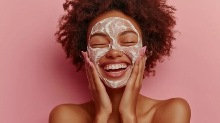 A happy woman with cream/soap, isolated on a pastel pink background. A beauty portrait of a smiling African American girl, smiling while washing her face during her morning skincare routine. 