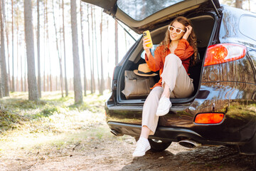 A young woman enjoys a sunny day by the car while using her smartphone, wearing stylish sunglasses and a bright orange blouse. Travel, weekend, nature, relax and lifestyle concept