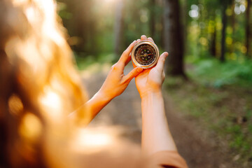 A hiker with curly hair holds a compass in their hands, carefully navigating through a sunlit forest filled with tall trees, greenery, and warm afternoon light.