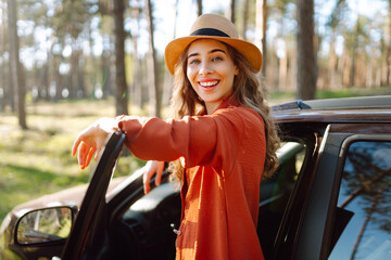 Wall Mural - Young woman enjoying a sunny day by her car in a serene forest setting during the afternoon. Summer trip. Lifestyle, travel, tourism, nature, active life.