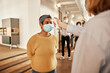 © Flamingo Images - Young female entrepreneur in a yellow shirt, having her temperature taken, by a colleague in an office community before she is allowed to enter the workplace. Her colleagues are queuing up behind her
