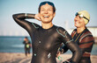 © Flamingo Images - Woman helping a laughing friend on a wetsuit for an open water swim. They have diving goggles on their foreheads and the sun hits their faces