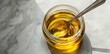 © Makki - A diagonal view of a glass jar with golden oil and a spoon on a white marble table, shown closeup.