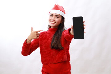 Wall Mural - Girl in christmas red turtleneck and santa claus hat shows blank smartphone screen on white isolated background, woman pointing advertises phone display in christmas clothes