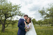 © Vasil - A bride and groom are standing in a field with trees in the background. The bride is holding a bouquet and the groom is holding her. Scene is happy and romantic