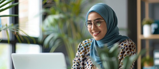 Leinwandbilder - A hijab clad woman in an office setting gazes happily at her work on a laptop her expression cheerful The scene is perfect for a copy space image