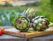 © coffeeflavour - Close-up of three artichokes on a wooden chopping board. Blurred background of the countryside.