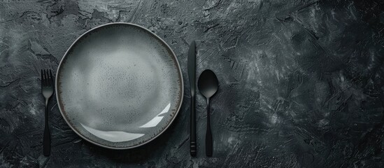 Poster - Dark textured concrete background with a round ceramic plate and cutlery setting the scene for dinner preparation with copy space image