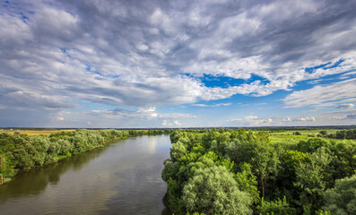 Wall Mural - A tranquil river meanders through vibrant greenery, reflecting a stunning sky filled with clouds, creating a peaceful atmosphere in the early afternoon light.