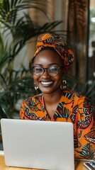 Wall Mural - Vibrant African-inspired fashion meets modern technology as a woman in colorful attire and headwrap smiles brightly while working on her laptop in a lush, plant-filled setting.