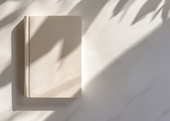 Photo of a minimalist blank journal on an elegant white table with sun rays and shadows. Top View of personal planner  Mockup paper notebook design with leaves and natural sunlight