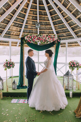 Wall Mural - A bride and groom stand under a green archway, holding hands. The bride is wearing a white dress and the groom is wearing a suit. The archway is decorated with flowers