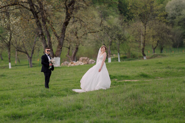 A bride and groom are standing in a grassy field. The bride is wearing a white dress and the groom is wearing a suit. They are both smiling and looking at the camera