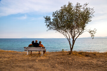 Idyllic landscape with people sitting on bench near tree in the background of a sea bay at sunset