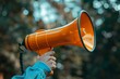 © raquel - Hand Holding a Bright Orange Megaphone Against a Blurred Natural Background