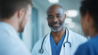 © Jane - smiling Black male doctor wearing glasses and white coat talking to two other doctors in hospital