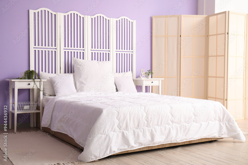 Interior of stylish bedroom with comfortable bed, white pillows, bedside tables and folding screen