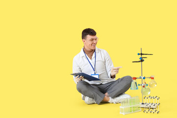 Wall Mural - Male chemist with clipboard pointing at equipment on yellow background