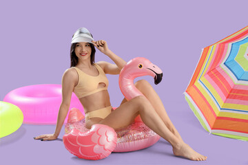 Poster - Beautiful young happy woman in swimsuit with inflatable rings and beach umbrella on purple background. Summer vacation concept