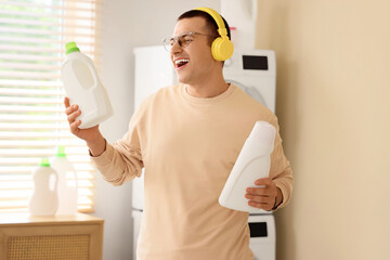 Canvas Print - Young man in headphones holding bottles of detergent in laundry room