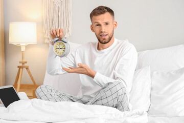 Wall Mural - Young man holding alarm clock on bed in bedroom. Insomnia concept