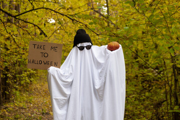 Man wearing ghost costume holding cardboard with text TAKE ME TO HALLOWEEN and pumpkin between orange yellow leaves in autumn forest for Halloween. Cool ghost creative holiday celebration fall