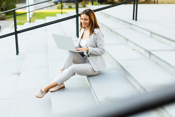 Business woman in a stylish outfit sits on outdoor steps while working on her laptop, enjoying a productive moment in a lively urban environment