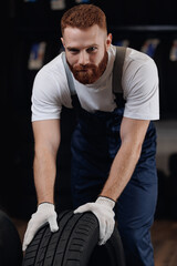 Wall Mural - Adult man mechanic in uniform rolls tire on black background