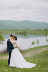 A bride and groom are standing in a grassy field by a lake, with the bride holding a bouquet. Concept of love and happiness, as the couple is celebrating their wedding day