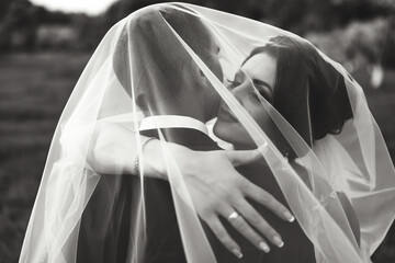 A bride and groom are embracing each other under a veil. The bride is wearing a white dress and the groom is wearing a suit. Scene is romantic and intimate