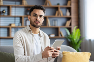 confident young man holding tablet in modern living room with plants and shelves, wearing casual att