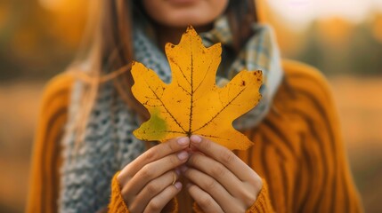 woman holding a yellow leaf in her hands, wearing autumn clothes, with a blurred outdoor background.