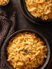 Two black bowls filled with creamy macaroni and cheese, garnished with rosemary, sit on a gray textured surface. Cheese slices on a wooden board are visible in the background a dark cloth.