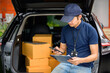 © เลิศลักษณ์ ทิพชัย - A delivery man stands at the back of a truck holding a clipboard and cardboard boxes, checking the address and cardboard boxes to confirm delivery