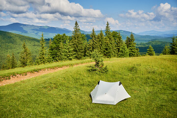 Wall Mural - White tourist tent on sunlit, grassy hilltop, surrounded by young pine trees. Panoramic view of lush, rolling mountains under bright blue sky with scattered clouds, creating idyllic camping spot.