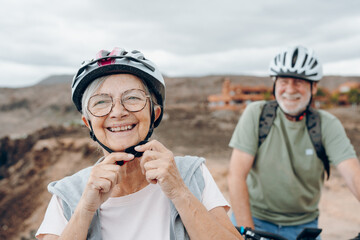 Canvas Print - Portrait of one old woman smiling and enjoying nature outdoors riding bike with her husband laughing. Headshot of mature female with glasses feeling healthy. Senior putting on helmet to go trip