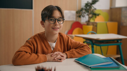 Wall Mural - Portrait little smiling Caucasian boy in eyeglasses healthy son looking at camera sitting at table primary school classroom smile kid schoolboy child baby pupil student studying education at desk