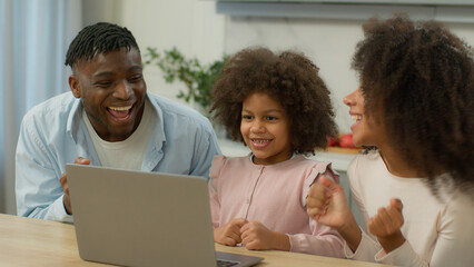 Happy family African American mother father with little daughter win laptop using pc celebrate achieve food delivery order discount dad mom with child kid baby girl computer victory at kitchen home