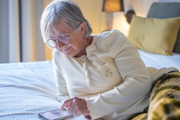 Wall Mural - Smiling senior woman lying in bedroom at home or hotel room looking at mobile phone enjoying tech and social. Elderly female online typing on smart phone