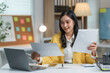 © amnaj - Asian businesswoman smiles while reviewing paperwork at her desk in her bright, modern office